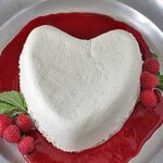 A heart-shaped white cheesecake sits on a metal platter, surrounded by raspberry sauce and garnished with fresh raspberries and mint leaves.