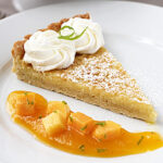 A slice of pie topped with whipped cream and a green garnish, dusted with powdered sugar, served on a white plate with mango cubes and sauce.