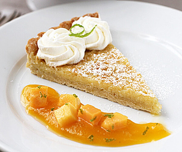 A slice of pie topped with whipped cream and a green garnish, dusted with powdered sugar, served on a white plate with mango cubes and sauce.