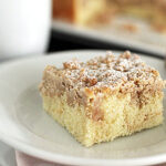 A square slice of crumb cake with a cinnamon crumb topping sits on a white plate next to a fork, with a blurred mug and more cake in the background.