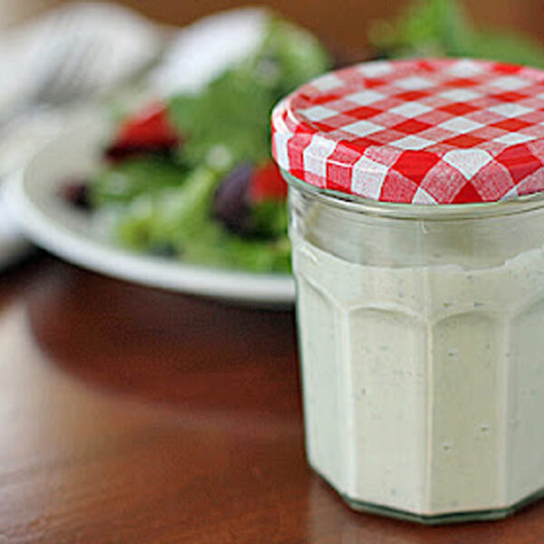 A glass jar with a red and white checkered lid filled with creamy dressing sits on a wooden table, with a plate of salad and a fork in the blurred background.