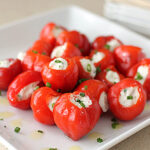 A white plate with bright red cherry peppers stuffed with creamy white cheese, garnished with chopped green herbs and a drizzle of olive oil. The peppers are arranged in a pile, ready to be served as appetizers.
