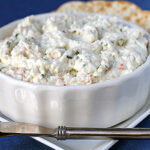 A white bowl filled with a creamy cheese spread sits on a square plate with round crackers in the background and a silver spreader in front of the bowl.