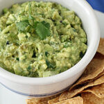 A bowl of chunky guacamole topped with a sprig of cilantro sits on a white plate next to a pile of crispy tortilla chips.