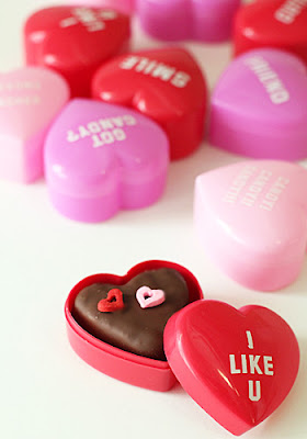 A red heart-shaped box with I LIKE U holds a chocolate treat topped with two small heart decorations. Several other pink and red heart-shaped boxes are scattered in the background.