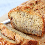 A loaf of banana bread on a wooden surface, with two slices cut in front of it. The bread has a golden-brown crust and a moist, textured interior. A knife is partially visible in the background.