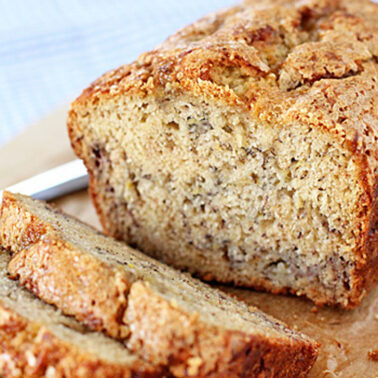 A loaf of banana bread on a wooden surface, with two slices cut in front of it. The bread has a golden-brown crust and a moist, textured interior. A knife is partially visible in the background.