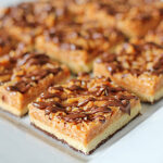 A plate of caramel shortbread bars topped with chopped nuts and drizzled with chocolate, arranged in neat rows on a white rectangular platter.