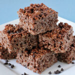 Six chocolate rice crispy treats are stacked on a white plate with a few small chocolate chips scattered around, against a light blue background.