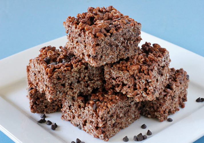 Six chocolate rice crispy treats are stacked on a white plate with a few small chocolate chips scattered around, against a light blue background.