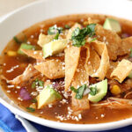 A bowl of tortilla soup topped with crispy tortilla strips, diced avocado, shredded chicken, chopped cilantro, corn, and crumbled cheese, served on a white plate with a blue napkin and spoon.