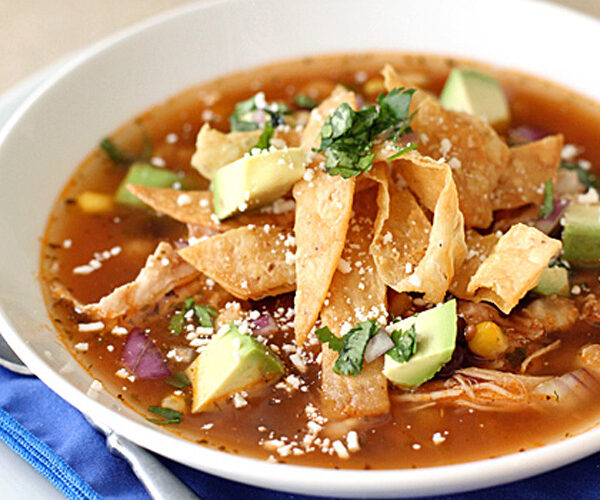 A bowl of tortilla soup topped with crispy tortilla strips, diced avocado, shredded chicken, chopped cilantro, corn, and crumbled cheese, served on a white plate with a blue napkin and spoon.