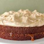A chocolate cake with a thick layer of caramel filling between the layers and topped with swirls of light brown frosting, displayed on a white plate against a pale green background.