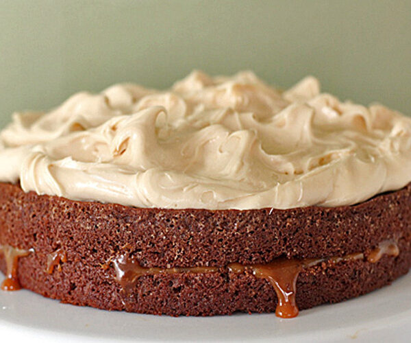 A chocolate cake with a thick layer of caramel filling between the layers and topped with swirls of light brown frosting, displayed on a white plate against a pale green background.