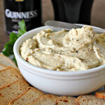 A white bowl of creamy dip sprinkled with pepper sits beside several slices of brown bread, with a can and a pint of Guinness beer in the background.