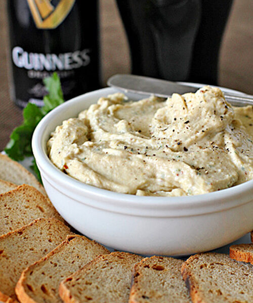 A white bowl of creamy dip sprinkled with pepper sits beside several slices of brown bread, with a can and a pint of Guinness beer in the background.