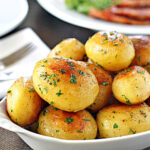 A white dish filled with golden, roasted baby potatoes garnished with chopped parsley. A plate, utensils, and another dish are blurred in the background.