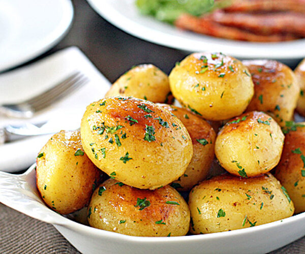 A white dish filled with golden, roasted baby potatoes garnished with chopped parsley. A plate, utensils, and another dish are blurred in the background.