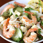 A white bowl filled with shrimp salad, including cucumbers, onions, parsley, cheese cubes, and lemon slices, set on a brown table with a fork partially visible.