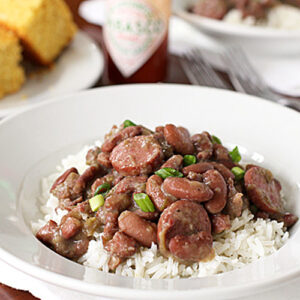A white bowl filled with white rice, topped with red beans and sausage slices, garnished with green onions. In the background, there is cornbread, a bottle of Tabasco sauce, and a fork and knife.