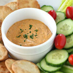 A bowl of creamy dip garnished with herbs is surrounded by pita chips, cucumber slices, cherry tomatoes, celery sticks, and lettuce on a white plate.
