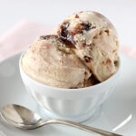 Two scoops of creamy ice cream with a swirl of chocolate in a white bowl, placed on a white saucer with a silver spoon beside it.