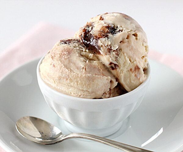 Two scoops of creamy ice cream with a swirl of chocolate in a white bowl, placed on a white saucer with a silver spoon beside it.