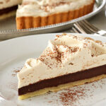 A slice of chocolate cream pie with a thick layer of whipped topping and cocoa powder sprinkled on top, served on a white plate. The whole pie is visible in the background.