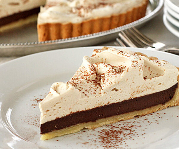 A slice of chocolate cream pie with a thick layer of whipped topping and cocoa powder sprinkled on top, served on a white plate. The whole pie is visible in the background.