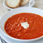 A white bowl filled with tomato soup, topped with a dollop of cream, is placed on a plate next to a spoon. In the background, slices of bread are visible on a separate plate.