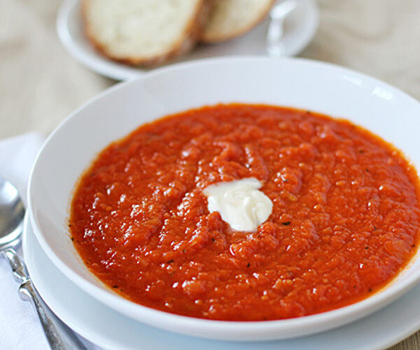 A white bowl filled with tomato soup, topped with a dollop of cream, is placed on a plate next to a spoon. In the background, slices of bread are visible on a separate plate.