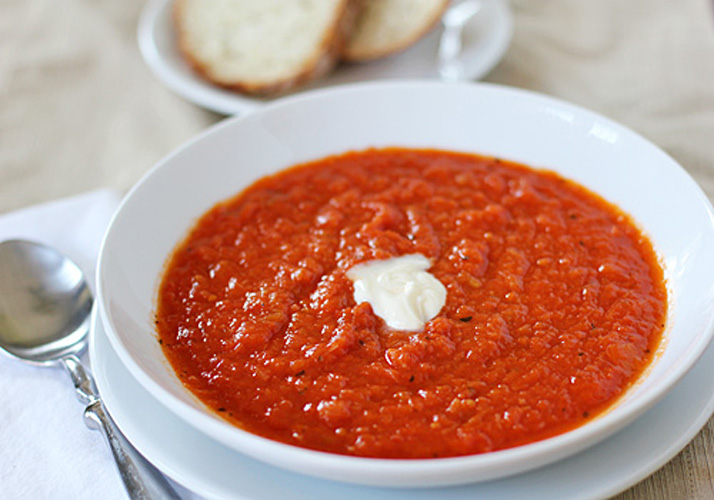 A white bowl filled with tomato soup, topped with a dollop of cream, is placed on a plate next to a spoon. In the background, slices of bread are visible on a separate plate.