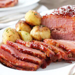 Sliced glazed corned beef with a side of seasoned roasted potatoes on a white plate, served with silverware, and a blurred background plate visible.