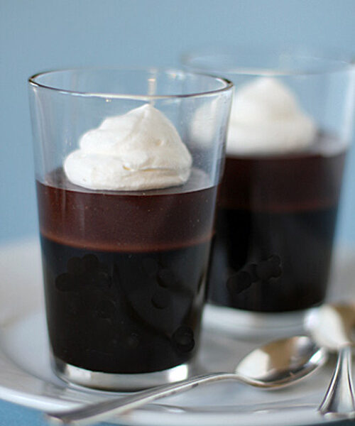 Two clear glasses filled with dark chocolate mousse, each topped with a swirl of whipped cream, are placed on a white plate next to two metal spoons against a light blue background.