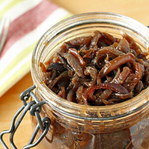 A glass jar filled with caramelized onion jam sits open on a wooden surface, with a striped cloth and a fork nearby in the background.