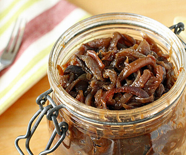A glass jar filled with caramelized onion jam sits open on a wooden surface, with a striped cloth and a fork nearby in the background.