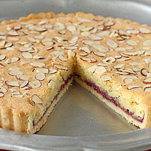 A baked tart with a golden crust, topped with sliced almonds. A slice has been removed, revealing a red jam layer between the crust and almond-flavored cake—just like a classic Raspberry Bakewell Tart recipe. The tart sits on a silver tray.