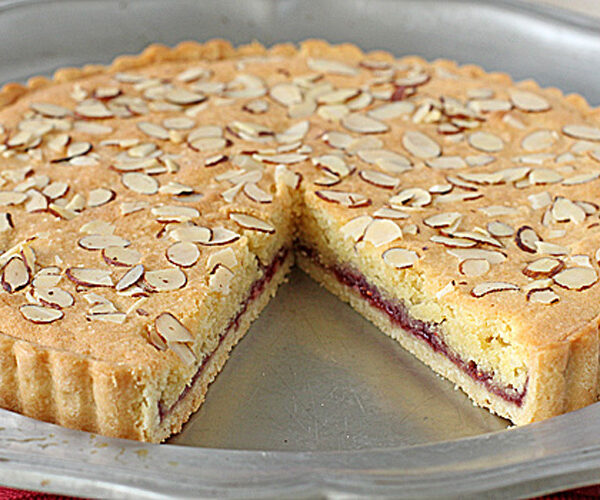 A baked tart with a golden crust, topped with sliced almonds. A slice has been removed, revealing a red jam layer between the crust and almond-flavored cake—just like a classic Raspberry Bakewell Tart recipe. The tart sits on a silver tray.