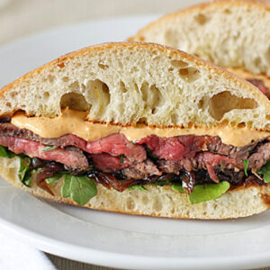 A steak sandwich with leafy greens, caramelized onions, and creamy sauce on ciabatta bread, sliced in half and served on a white plate.