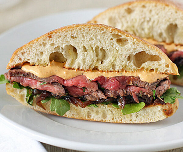 A steak sandwich with leafy greens, caramelized onions, and creamy sauce on ciabatta bread, sliced in half and served on a white plate.