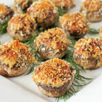 A white plate displays several stuffed mushrooms topped with golden-brown breadcrumbs, arranged on a bed of fresh dill.