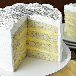 A tall, four-layer poppy seed cake with white frosting and a creamy yellow filling, topped with sprinkled poppy seeds. A slice is served on a plate in the background next to forks.