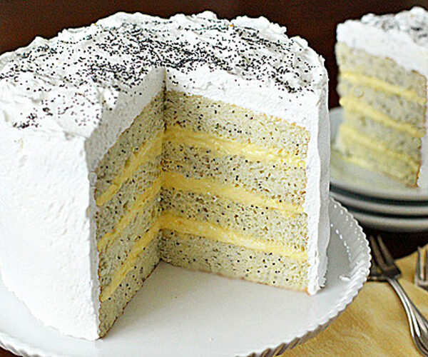 A tall, four-layer poppy seed cake with white frosting and a creamy yellow filling, topped with sprinkled poppy seeds. A slice is served on a plate in the background next to forks.