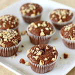 Seven chocolate muffins topped with chopped nuts are arranged on a white square plate, with a few nut pieces scattered around the muffins.