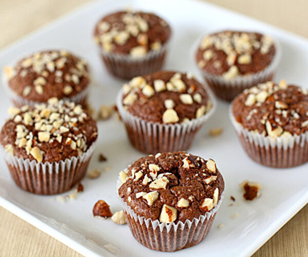 Seven chocolate muffins topped with chopped nuts are arranged on a white square plate, with a few nut pieces scattered around the muffins.
