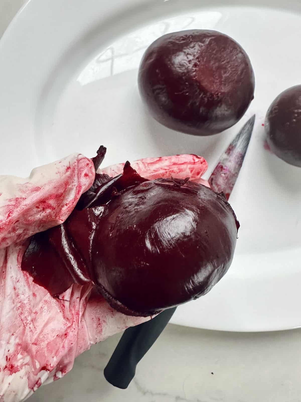 A gloved hand holds a peeled beet with deep red juice staining the glove. Two more peeled beets and a knife with beet juice are on a white plate in the background.