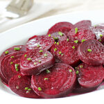 Sliced cooked beets arranged on a white oval plate, garnished with finely chopped green herbs, with a serving fork and spoon in the background.