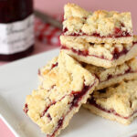 A plate stacked with crumbly jam bars, featuring a golden crust and a layer of red fruit filling, with a jar of jam in the background on a pink surface.
