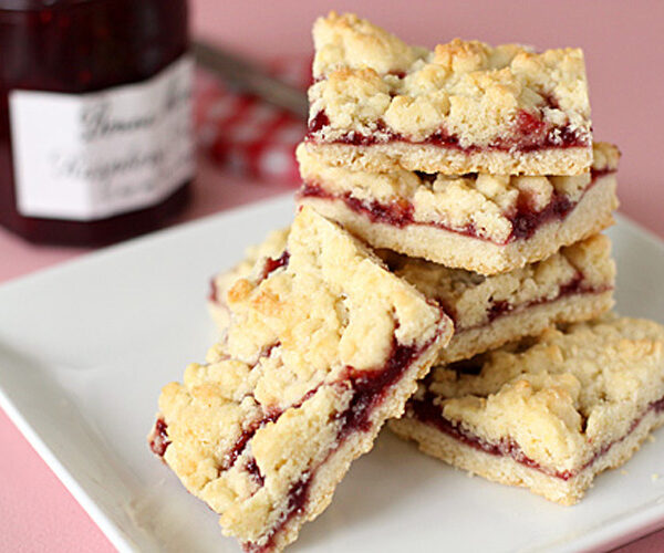 A plate stacked with crumbly jam bars, featuring a golden crust and a layer of red fruit filling, with a jar of jam in the background on a pink surface.