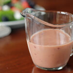 A glass pitcher filled with creamy pink salad dressing sits on a wooden table, with a blurred plate of salad in the background.